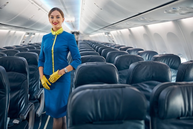 Mezzo busto ritratto di donna allegra felice in tuta blu assistente di volo in piedi sul bordo di un aereo commerciale vuoto