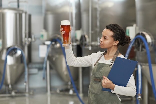 Mezzo busto ritratto di giovane donna afro-americana che tiene in mano un bicchiere di birra mentre ispeziona la qualità della produzione presso la fabbrica di birra, copia spazio