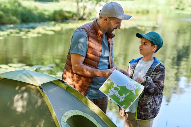 Mezzo busto ritratto di padre e figlio guardando la mappa mentre vi godete il campeggio insieme sul lago, copia dello spazio