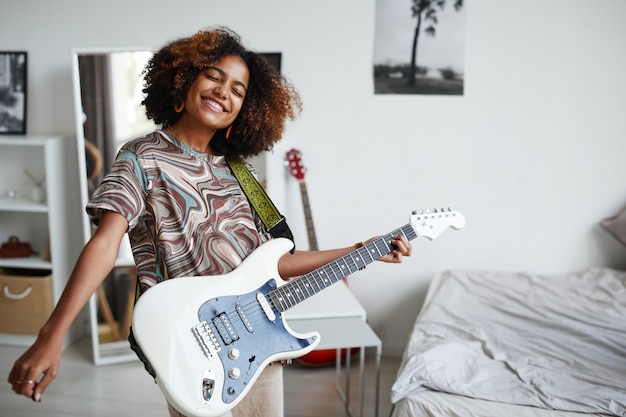 Mezzo busto ritratto di un'adolescente afro-americana sorridente che suona la chitarra elettrica a casa, copia spazio