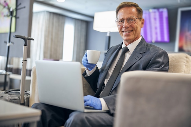 Mezzo busto ritratto di un elegante uomo d'affari sorridente che usa il taccuino mentre beve caffè con guanti in lattice in aeroporto