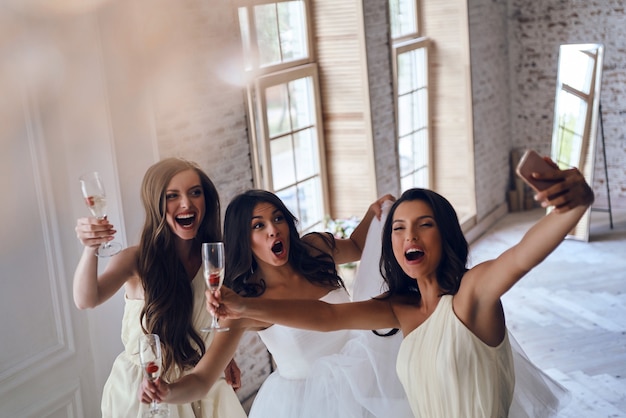 Mi sento così felice! Vista dall'alto di una giovane sposa attraente e due damigelle che fanno smorfie mentre si fanno selfie nel camerino