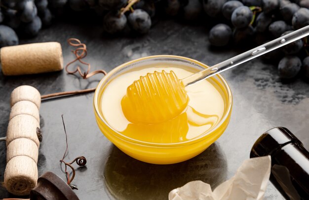 Miele in vaso di vetro con cucchiaio di miele su tavola di pietra scura con cavatappi vino formaggio Uva nera. Degustazione di vini con snack di formaggio al vino rosso miele su sfondo di cemento scuro.