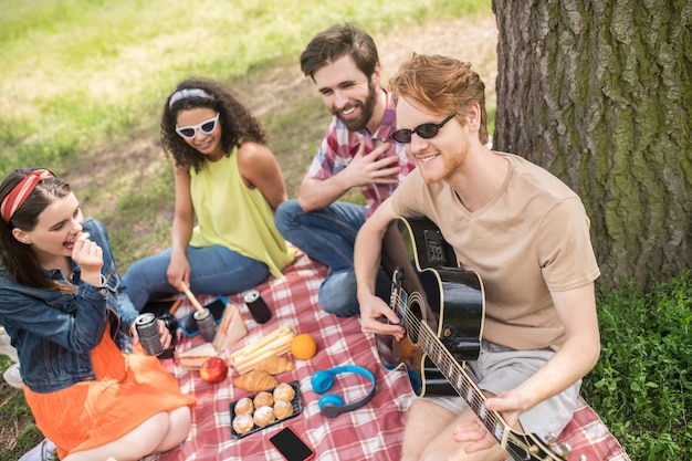 Miglior tempo. Felici ragazze e ragazzi che si divertono nel fine settimana nella natura suonando la chitarra mangiando comunicando