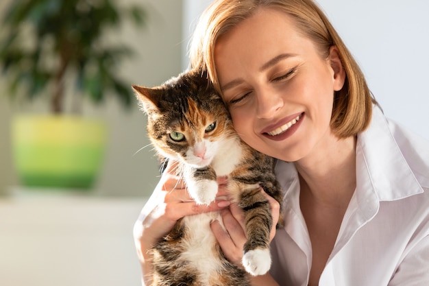 Miling donna in camicia bianca che abbraccia e abbraccia con tenerezza e amore gatto domestico in casa