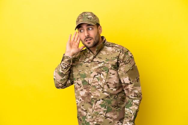 Militare isolato su sfondo giallo ascoltando qualcosa mettendo la mano sull'orecchio