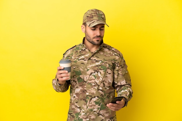 Militare isolato su sfondo giallo che tiene il caffè da portare via e un cellulare