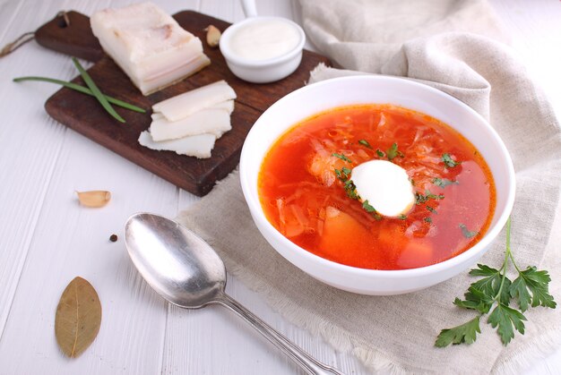 Minestra rossa del borscht in ciotola bianca con panna acida e prezzemolo, vista superiore, su fondo bianco di legno.