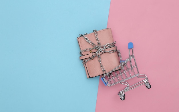 Mini carrello della spesa con portafoglio in pelle avvolto da catena in acciaio su fondo rosa azzurro. Vista dall'alto