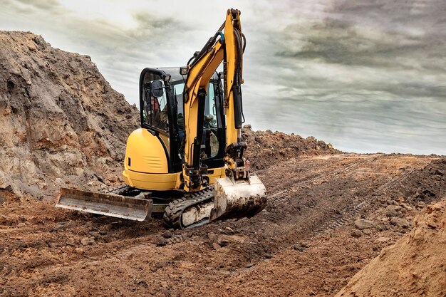 Mini escavatore in cantiere sul bordo di una fossa contro un cielo blu nuvoloso Attrezzatura da costruzione compatta per lavori di sterro Un assistente indispensabile per i lavori di sterro