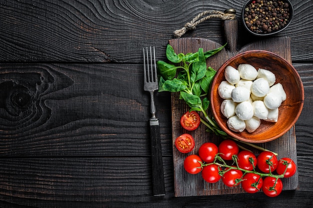 Mini palline di mozzarella italiana, basilico e pomodorini pronti per la cottura Insalata caprese. Fondo in legno nero. Vista dall'alto. Copia spazio.