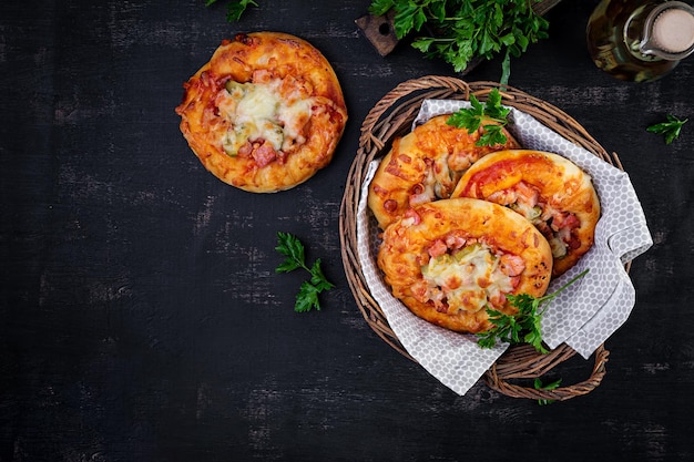 Mini pizza per bambini piccole torte aperte con prosciutto, salsa di pomodoro, mozzarella e formaggio in cesto su sfondo scuro Vista dall'alto