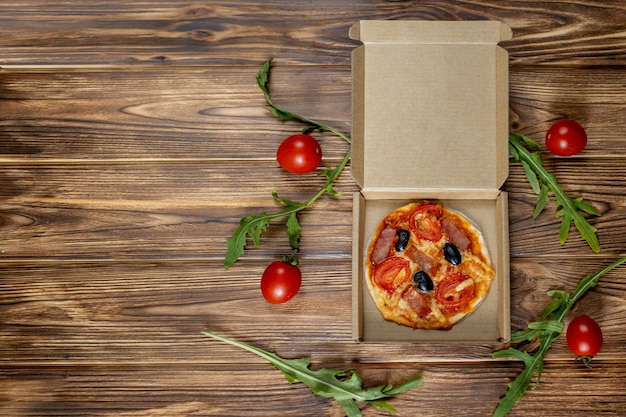 Mini pizza preparata da un bambino con pomodori, olive e prosciutto su fondo in legno.