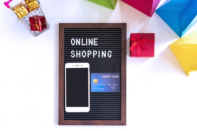 Miniatura dei contenitori di regalo in carrello, smartphone, parola sulla struttura nera, carta di credito e borse variopinte sullo scrittorio bianco. Concetto di shopping online, tecnologia e stile di vita.