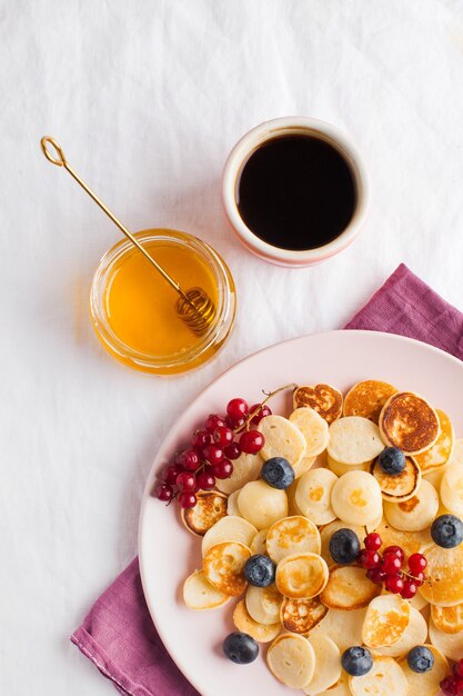 Minuscole frittelle con frutti di bosco, miele e caffè su una tovaglia bianca e spazio per il testo