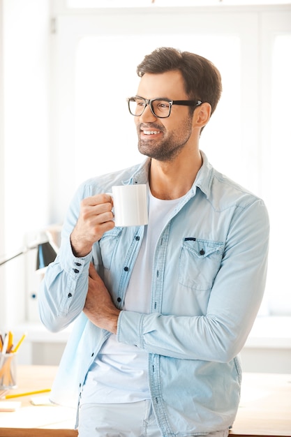 Minuto per il caffè fresco. Bel giovane con gli occhiali che tiene una tazza di caffè e guarda in modo mentre si trova contro il suo posto di lavoro