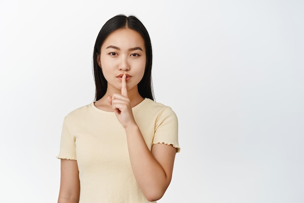 Misteriosa ragazza asiatica fa shh segno shush mostrando gesto tabù in piedi in maglietta gialla su sfondo bianco
