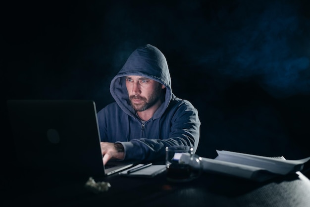 Misterioso uomo barbuto in un cappuccio che hackera una password su un laptop al buio