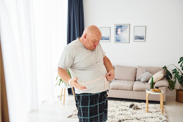 Misurare la vita L'uomo in sovrappeso divertente in abiti casual è in casa a casa