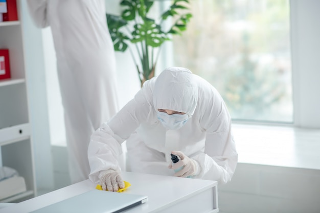 Misure protettive. Lavoratore medico in indumenti protettivi e tavolo disinfettante maschera medica, un altro lavoratore in piedi dietro