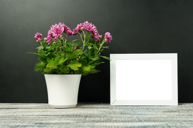 Mock up di una cornice bianca con un fiore in un vaso bianco su un tavolo in legno, muro nero. Orientamento verticale