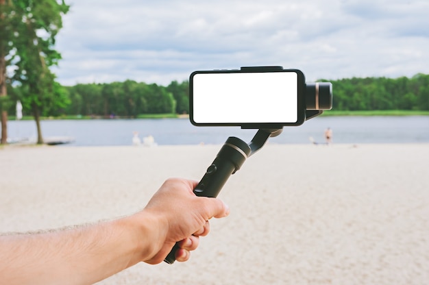 Mock up di uno smartphone con uno stabilizzatore della fotocamera nella mano di un uomo. Sullo sfondo di una spiaggia di sabbia e della natura con un lago.