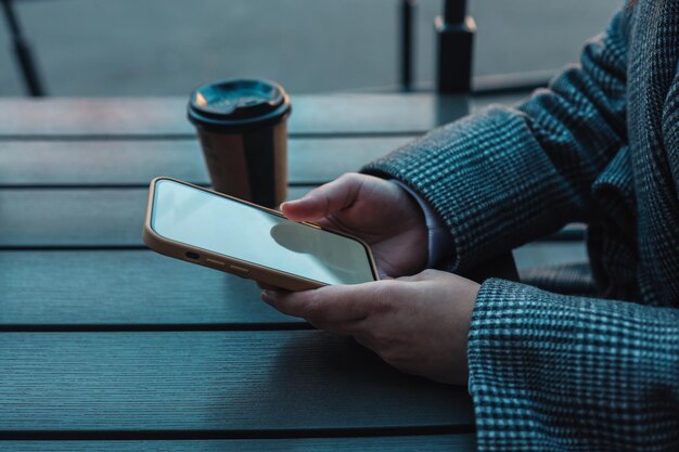 Mock up immagine di mani femminili che tengono il telefono cellulare con schermo vuoto e tazza di caffè in un bar per strada.