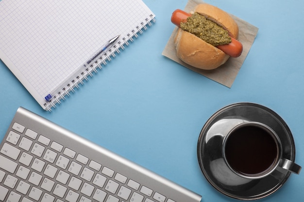 Mock-up sul posto di lavoro dell'ufficio - caffè, hot dog snack, quaderno, tastiera sul tavolo blu, vista piana, vista dall'alto