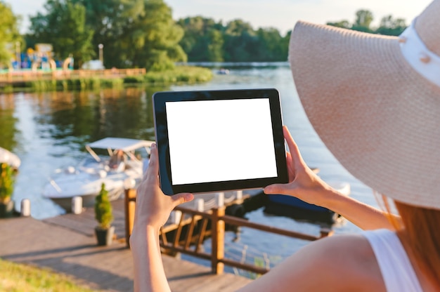 Mock up tablet nelle mani di una ragazza con un grande cappello. Sullo sfondo di un terrapieno in legno con acqua e una barca. Concetto di tecnologia e turismo.
