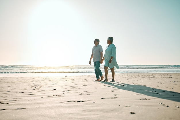 Mockup al tramonto e una coppia di anziani che camminano sulla spiaggia tenendosi per mano durante un appuntamento romantico insieme Ama la natura o il legame con un uomo e una donna anziani che fanno una passeggiata sulla costa durante l'estate