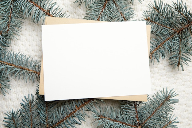 Mockup di biglietto di auguri di Natale con busta e rami di abete su sfondo pullover lavorato a maglia bianco vista dall'alto piatto