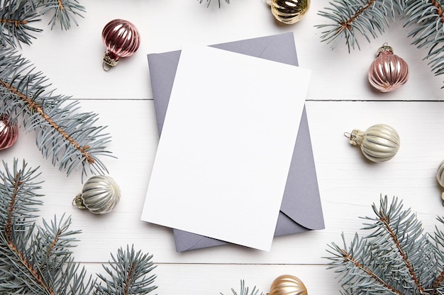 Mockup di biglietto di auguri di Natale con busta rami di abete verde e palline su fondo di legno bianco vista dall'alto piatto lay Mockup di biglietto per le vacanze invernali con decorazioni