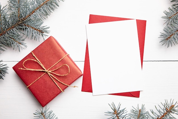 Mockup di biglietto di auguri di Natale con confezione regalo busta rossa e ramo di abete fresco su fondo di legno bianco vista dall'alto piatto lay Card vacanze invernali vuote