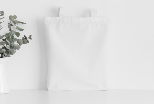 Mockup di borsa tote bianca con un eucalipto in un vaso su un tavolo bianco