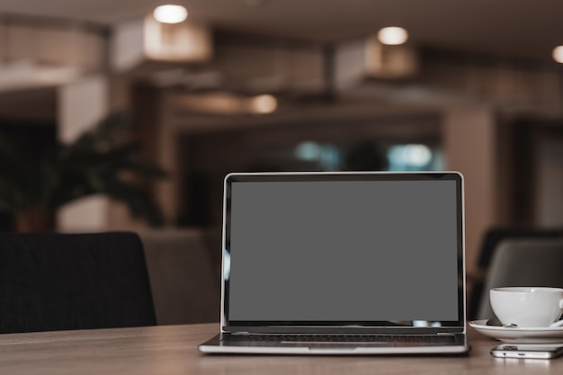 Mockup di computer portatile con schermo vuoto con tazza di caffè e smartphone sul tavolo dello sfondo della caffetteria, schermo grigio nero