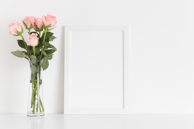 Mockup di cornice bianca con un bouquet di rose rosa in un vaso di vetro su un tavolo bianco Orientamento verticale