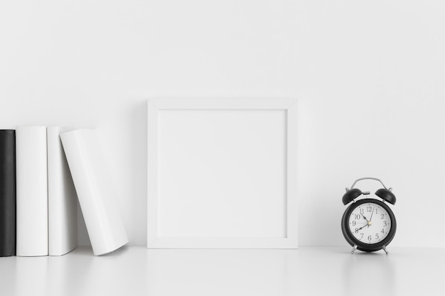 Mockup di cornice quadrata bianca con libri e un orologio su un tavolo bianco