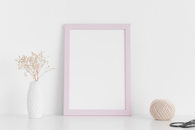 Mockup di cornice rosa con accessori per l'area di lavoro e gypsophila in un vaso su un tavolo biancoOrientamento verticale