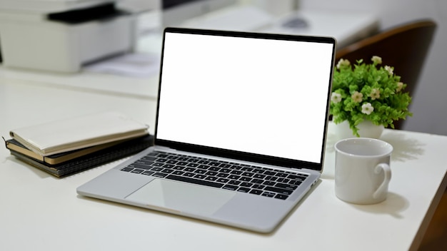 Mockup di schermo bianco per laptop aperto con un libro della tazza di caffè e una pianta decorativa sulla scrivania dell'ufficio