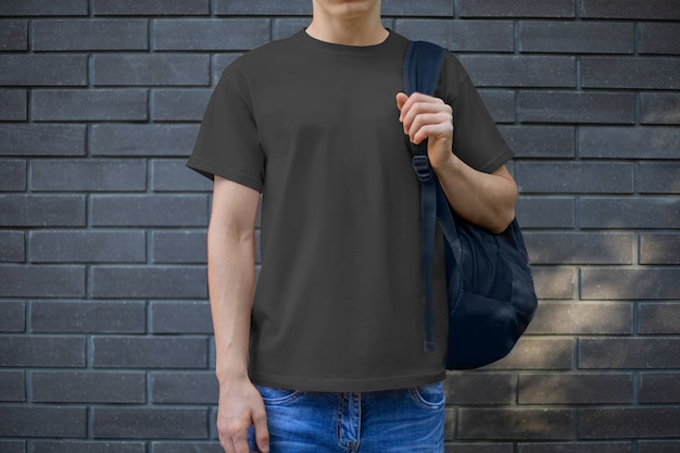 Mockup di una t-shirt da uomo nera su un giovane ragazzo contro un muro di mattoni, vista frontale. Un modello di abbigliamento casual per la presentazione del design e della pubblicità nel negozio online.