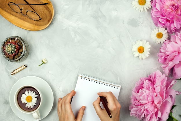 Mockup femminile flatlay con peonie rosa, margherite, bicchieri, tazza di tè caldo, cactus e mani femminili scrivendo nel quaderno su sfondo di cemento con copyspace