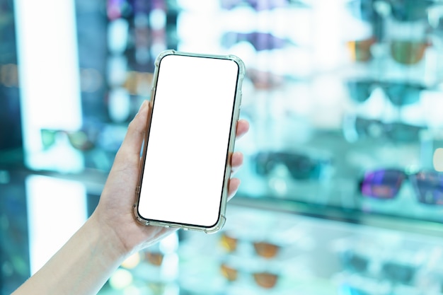 Mockup, mani che tengono il telefono cellulare con schermo bianco vuoto nel negozio di occhiali sfocati, concetto di pagamento digitale