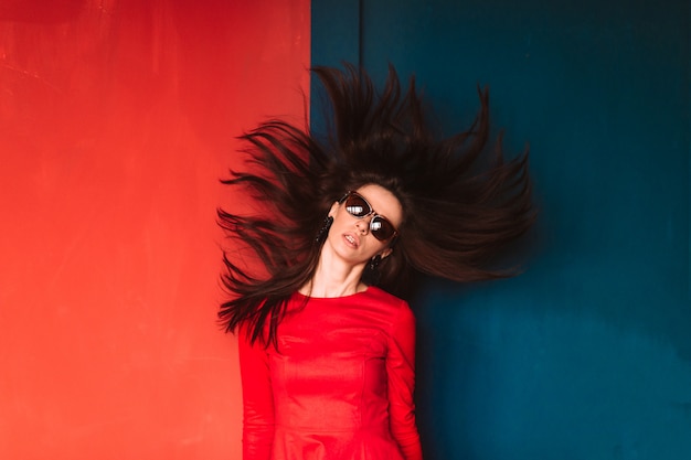 Moda bella ragazza con i capelli lunghi in occhiali da sole e abito rosso elegante in posa sul muro rosso blu. Elegante ragazza bruna con i capelli volanti. Concetto di acconciatura professionale