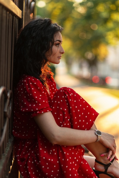 Moda bella ragazza italiana elegante solitaria con capelli ricci in un vestito rosso vintage alla moda si siede vicino a una recinzione in città al tramonto