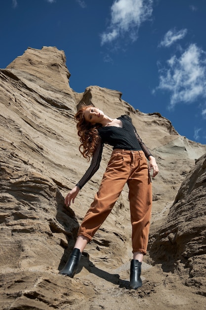 Moda donna dai capelli rossi in jeans arancioni in posa in natura vicino a rocce sabbiose