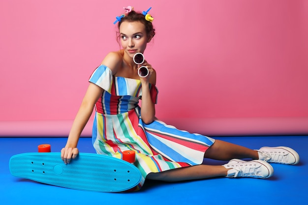 Moda donna fresca in posa con lo skateboard. Donna giovane hipster con bigodini tra i capelli, occhiali da sole, scarpe da ginnastica bianche. girato in studio