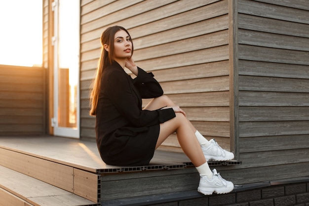 Moda donna in abiti alla moda con scarpe da ginnastica si siede vicino a una parete di legno al tramonto