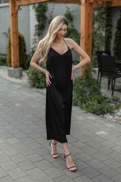 Moda donna piuttosto elegante e chic con i capelli biondi in elegante abito nero lungo sexy strappy con scarpe cammina per strada