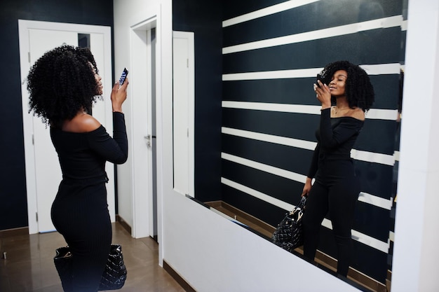 Moda giovane bella donna afroamericana con acconciatura afro e hangbag in posa indossare in nero elegante Fare foto allo specchio per telefono