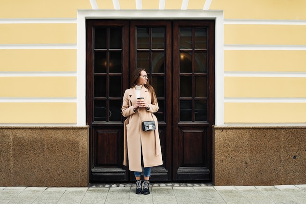 Moda giovane donna che cammina per strada, con un caffè per andare e un telefono cellulare